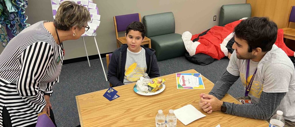 a women wearing a black and white striped blouse bending over to speak with a young boy who is sitting at a table with his reading mentor