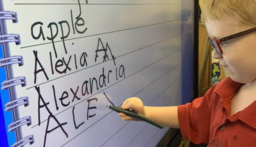 child in red shirt writing name on whiteboard